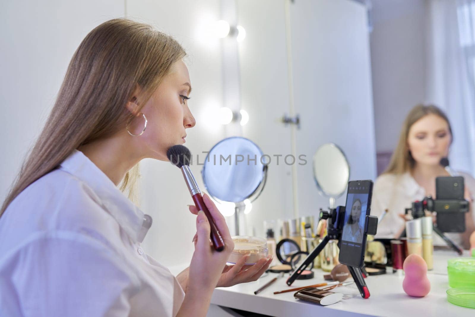 Young woman recording video, makeup beauty blog using smartphone. Female sitting in front of mirror, talking about cosmetics and showing application. Beauty, fashion, care, technology, youth concept