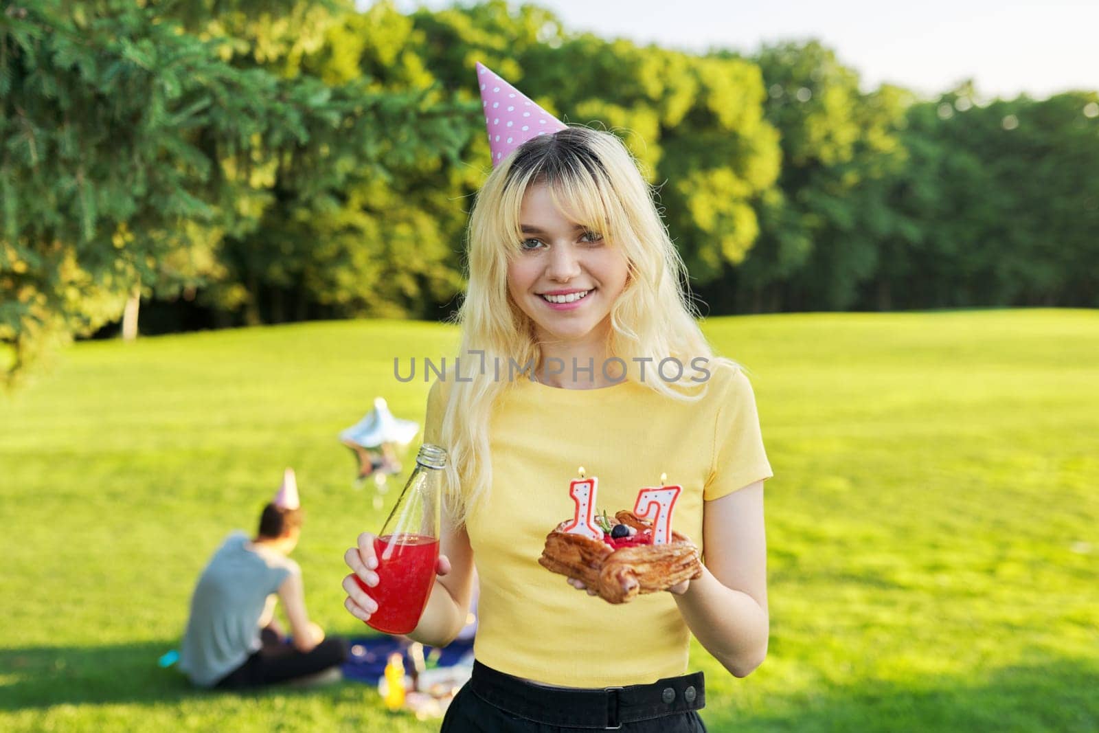 Beautiful teenage girl in festiv hat on birthday with cake and candles. Happy blonde female at outdoor picnic party holding cake with candles 17. Adolescence youth age beauty, holiday birthday concept