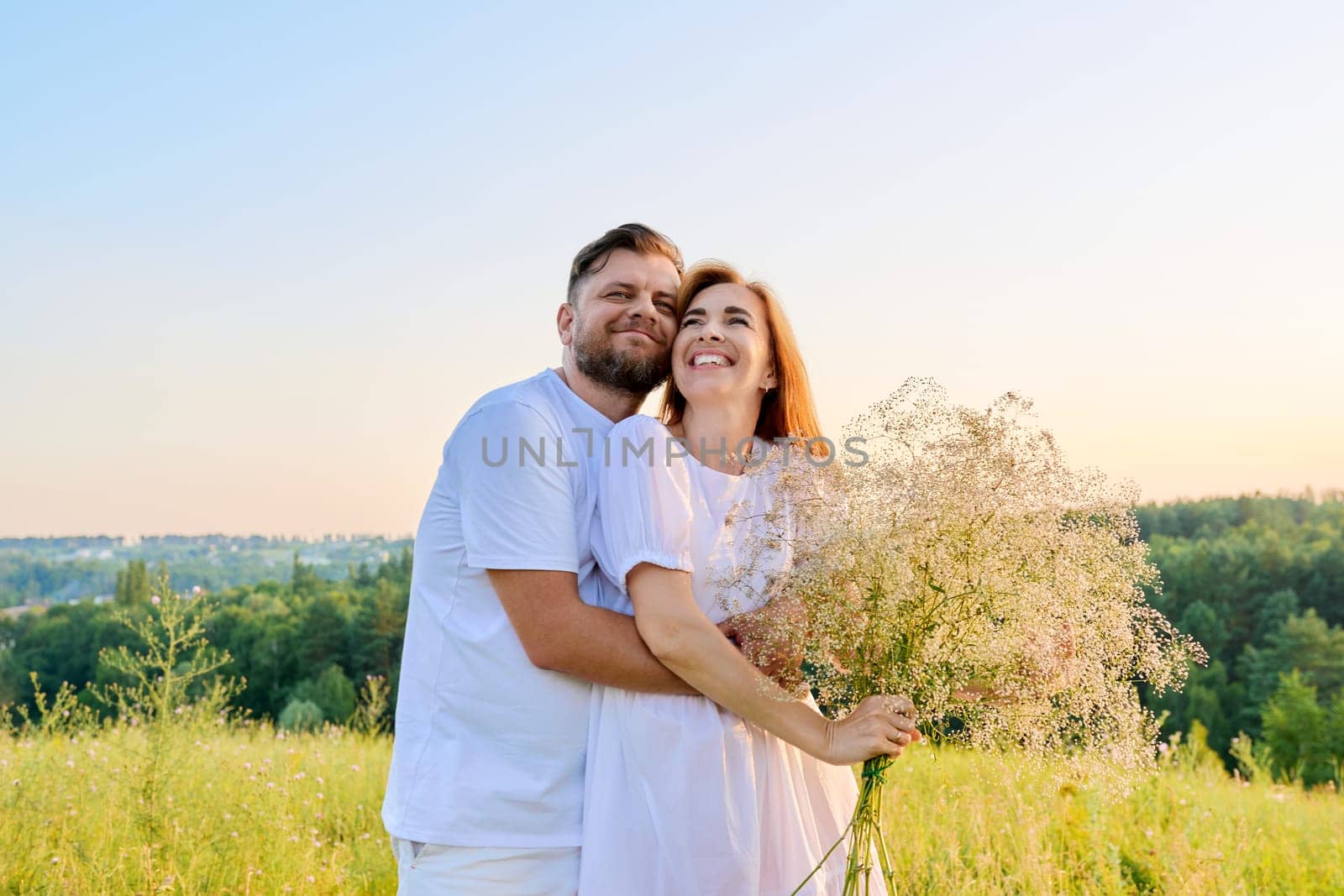 Love and romance of middle-aged couple. Happy hugging man and woman in white dress with bouquet of flowers, summer nature wild meadow. Relationship, holiday, date, wedding, family, people 40s concept