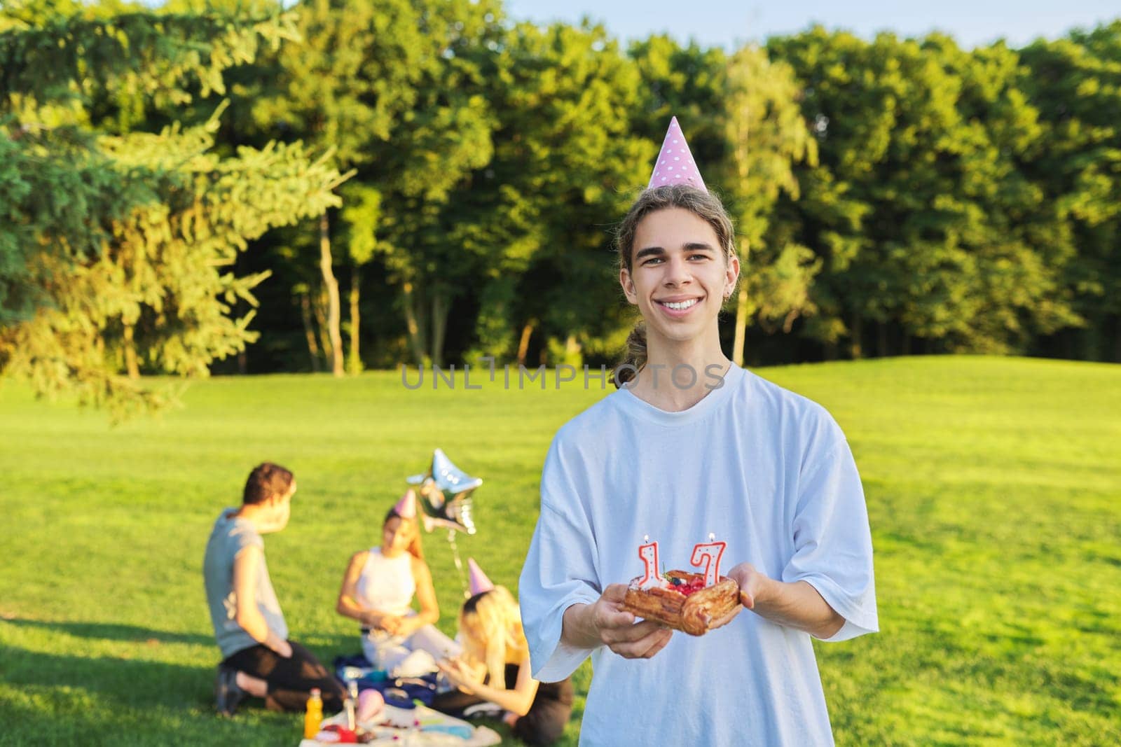 Happy guy teenager in birthday hat with cake with candles 17. Outdoor picnic party with friends in park on grass. Adolescence, having fun, holiday, celebration, birthday, age concept