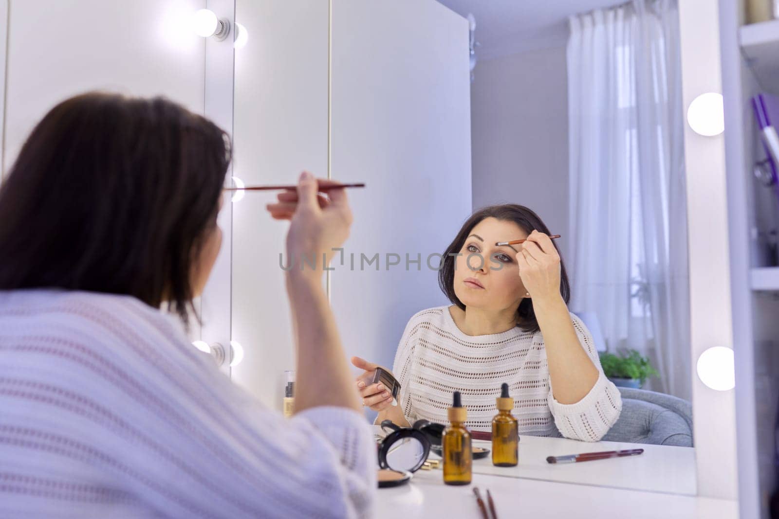 Beautiful middle aged woman doing makeup in front of a mirror. Cosmetics, beauty, make-up, face care, skin care, women 40s age concept