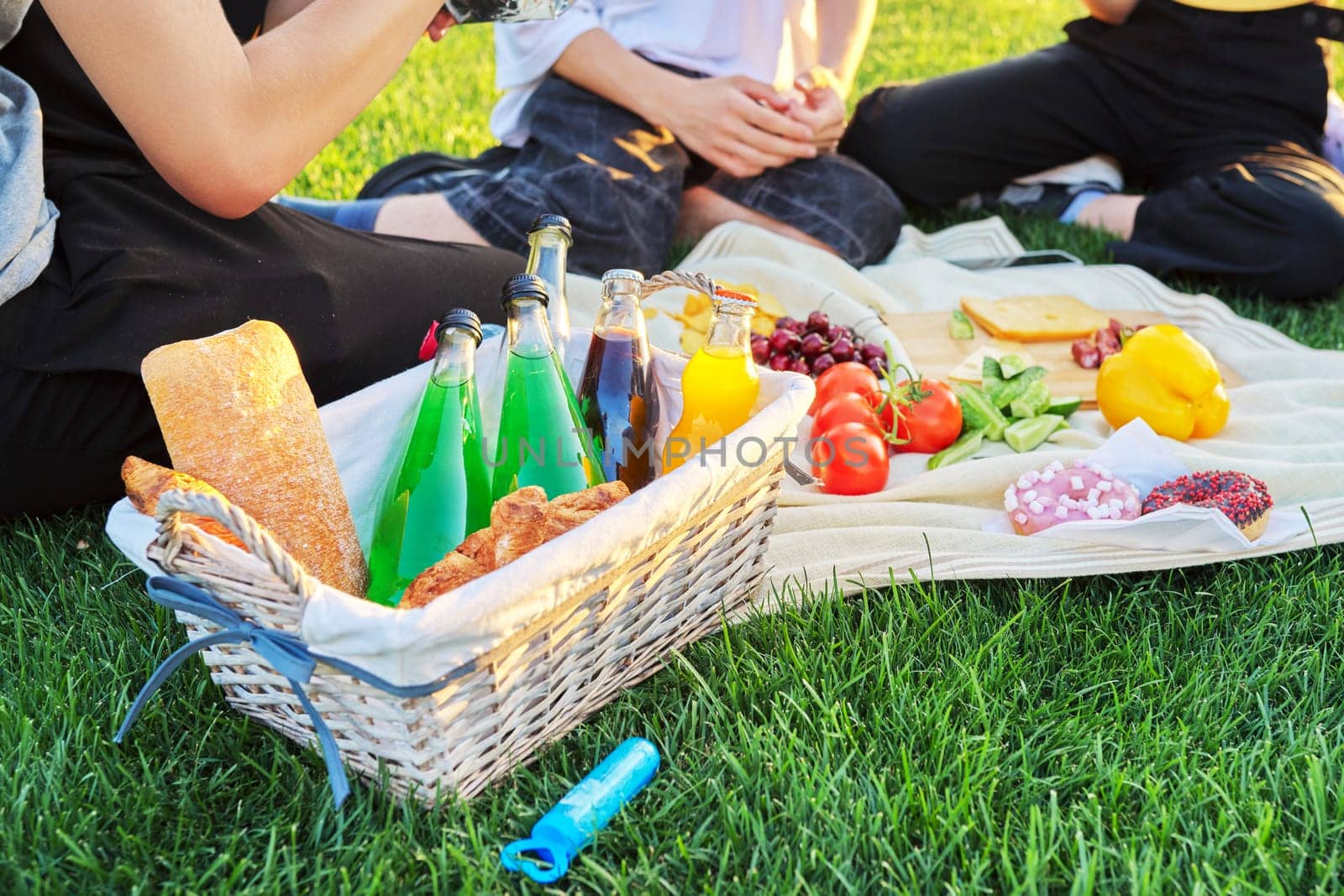 Close-up food and drink for picnic on green grass. Group of young people resting in nature, basket with bread, cheese fruits vegetables snacks juices on tablecloth. Youth, leisure, lifestyle, fun,