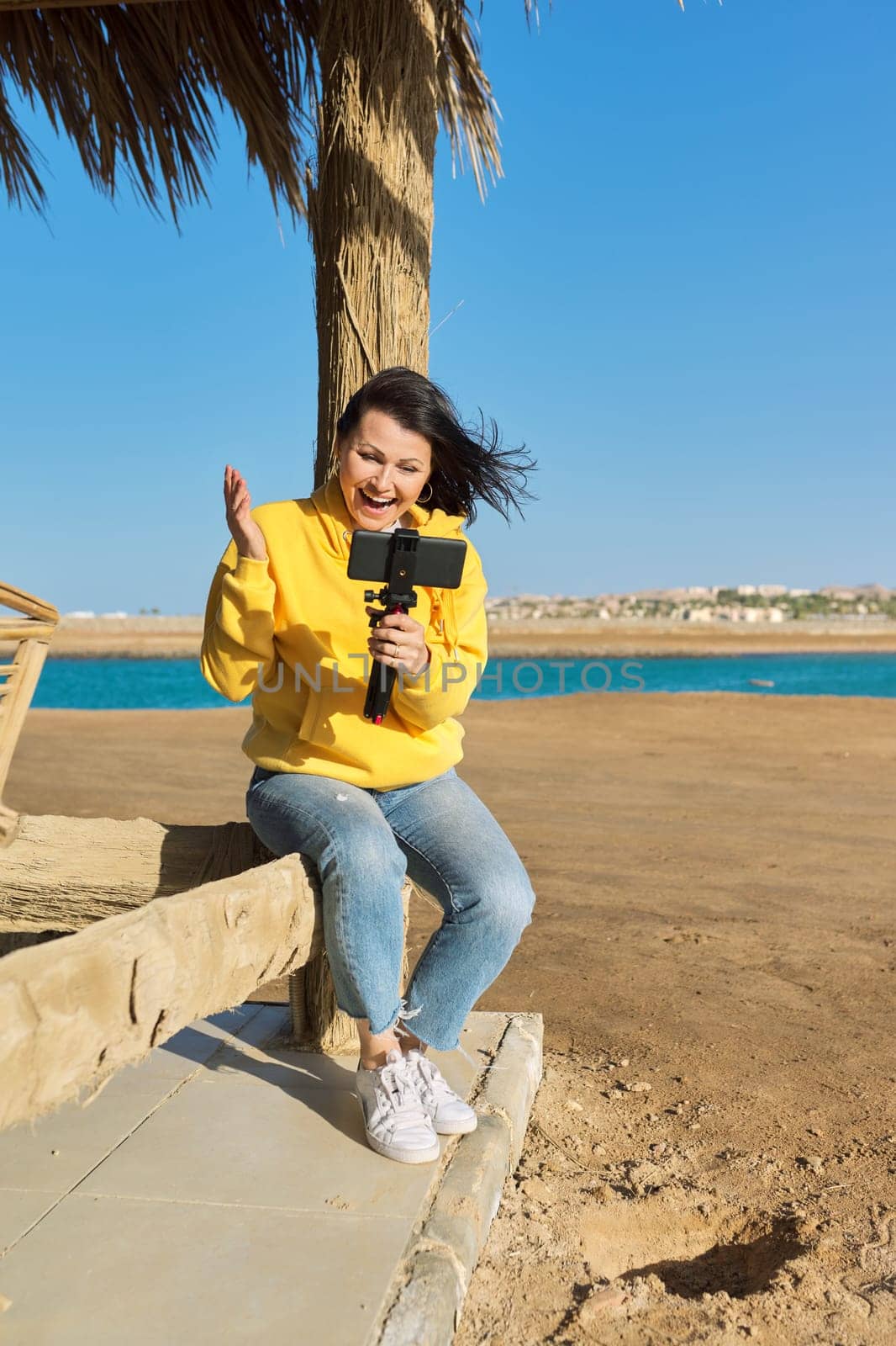 Middle aged woman on the beach talking online using smartphone. Video call, video meeting, chatting, tele technology concept