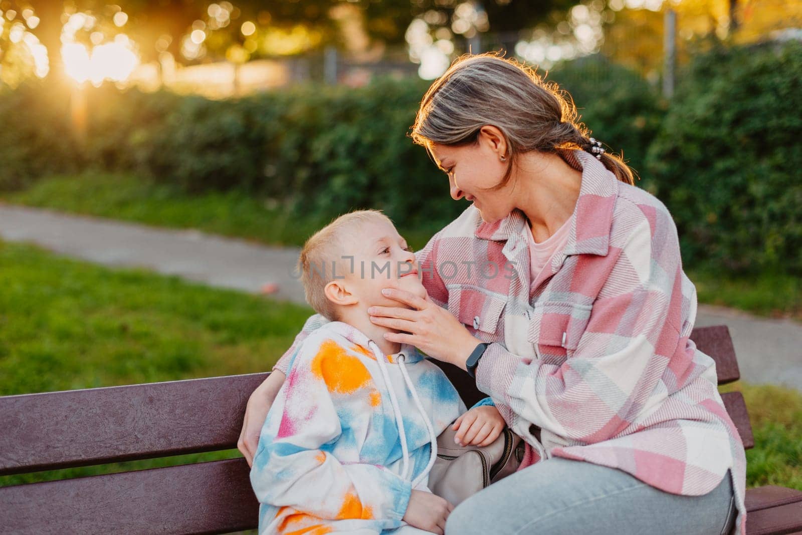 mother and son sit on a park bench in the rays of the setting sun. the concept of a family. Mother's Day. beautiful girl (mother) with a boy (son) in the park in the park are sitting on a bench at sunset by Andrii_Ko