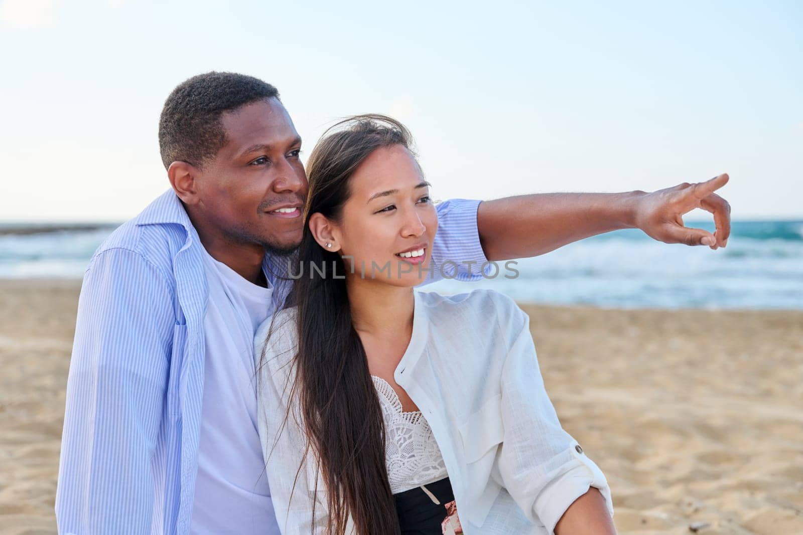 Happy multiethnic family resting on beach together. Asian woman and African man seated hugging each other on sand. Relationships, vacation travel and tourism at seaside resorts, people 20 30 years old