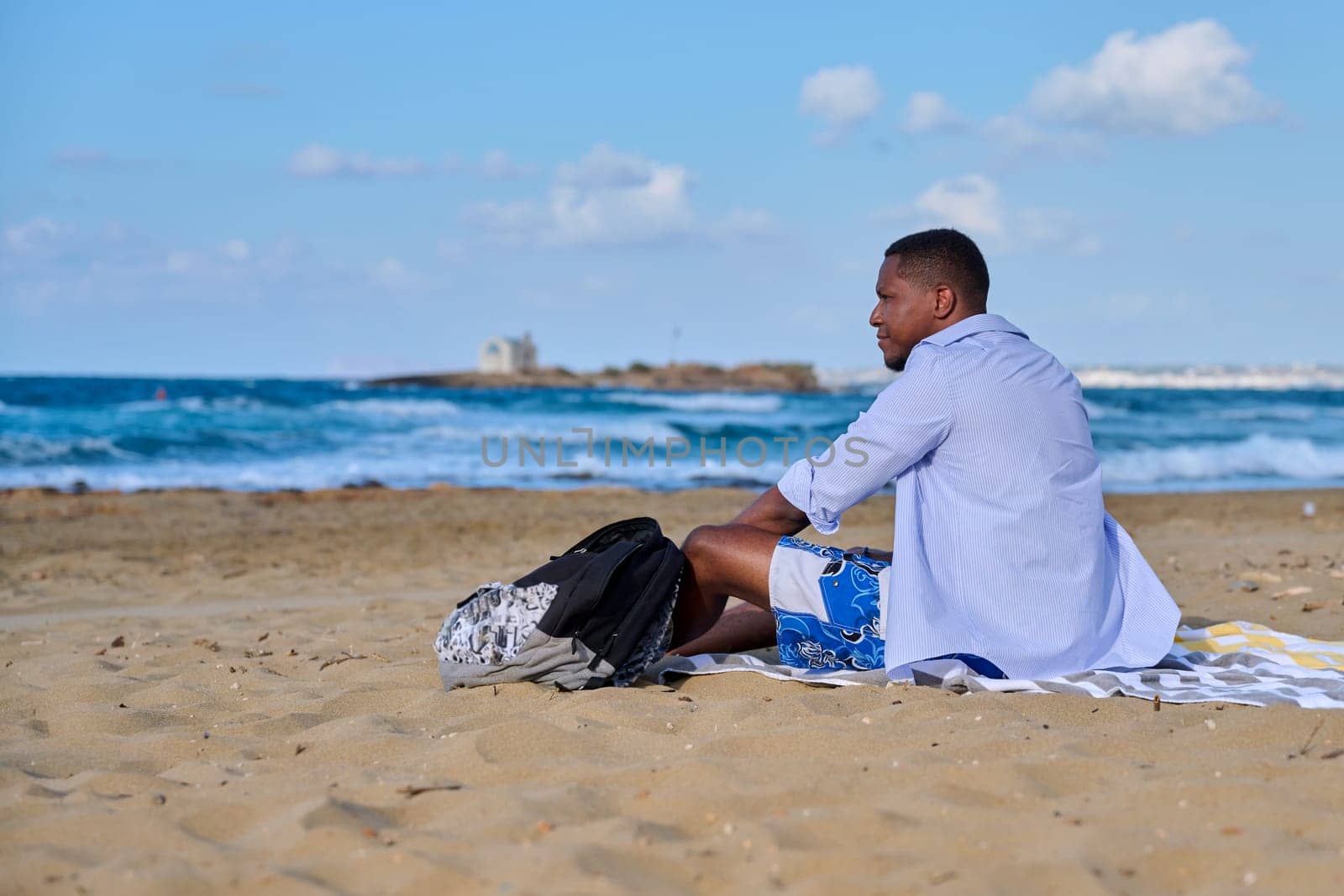 Young positive male tourist sitting resting on beach, copy space. Smiling African American male 30 years old sitting on sand, sea nature background. Lifestyle, travel, tourism, people concept