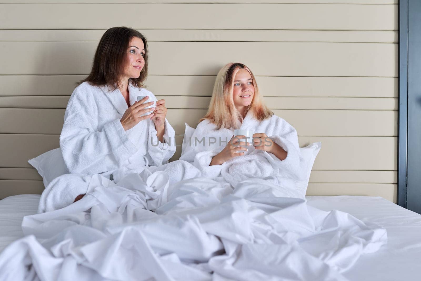 Happy mature mother and teenage daughter drinking coffee, talking, smiling. Women in white bathrobes, resting in bed, vacation together. Communication between parent and adolescent child, family