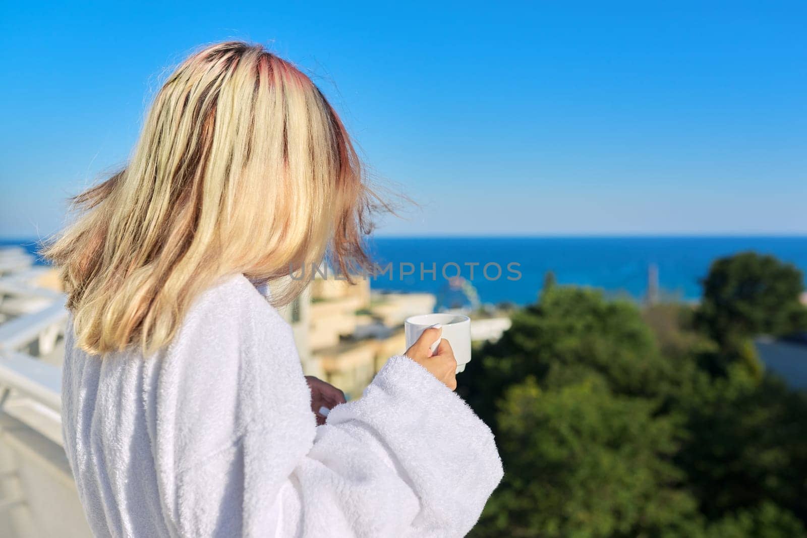 Summer vacation at seaside resort. Young woman in white bathrobe enjoying cup of coffee and sunny landscape of sea resort on balcony terrace of hotel, blue sky copy space, back view