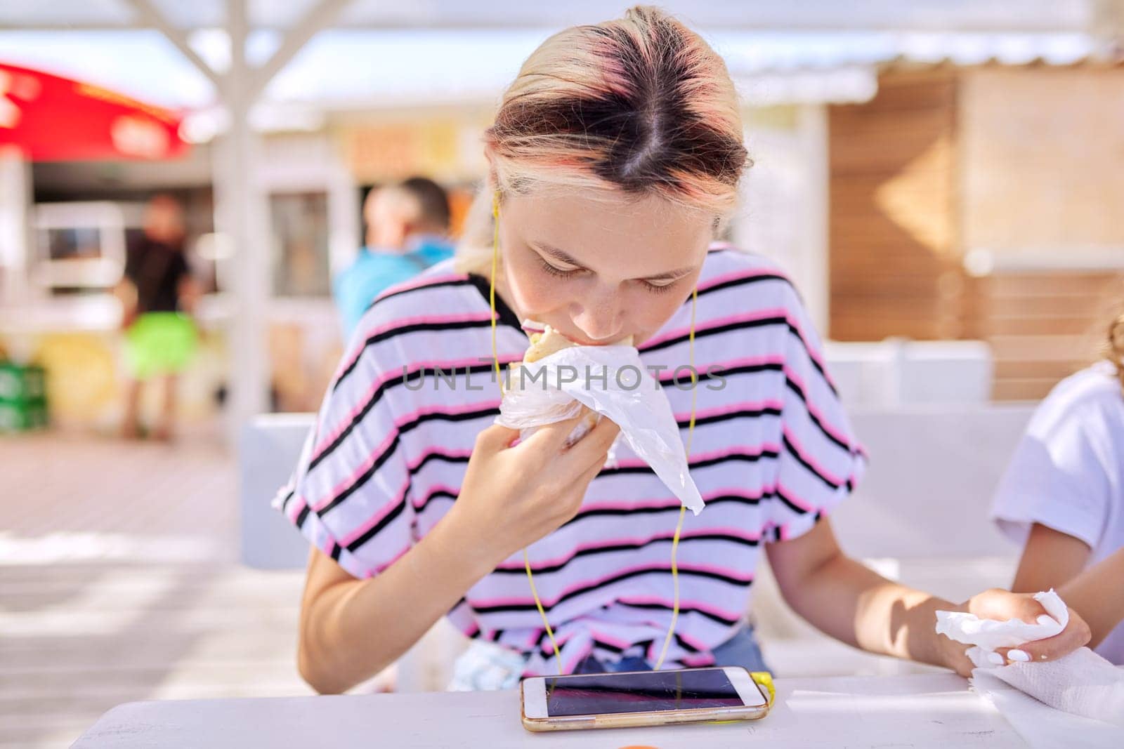 Fast food, summer, lifestyle, teenagers. Teen girl eating a sandwich, burger, looking at the smartphone screen in headphones