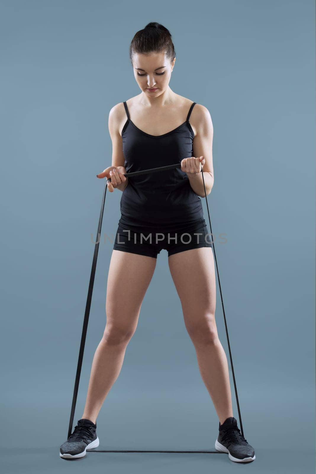 Young athletic woman doing arm and leg exercises using fitness elastic band with strong resistance, on gray studio background