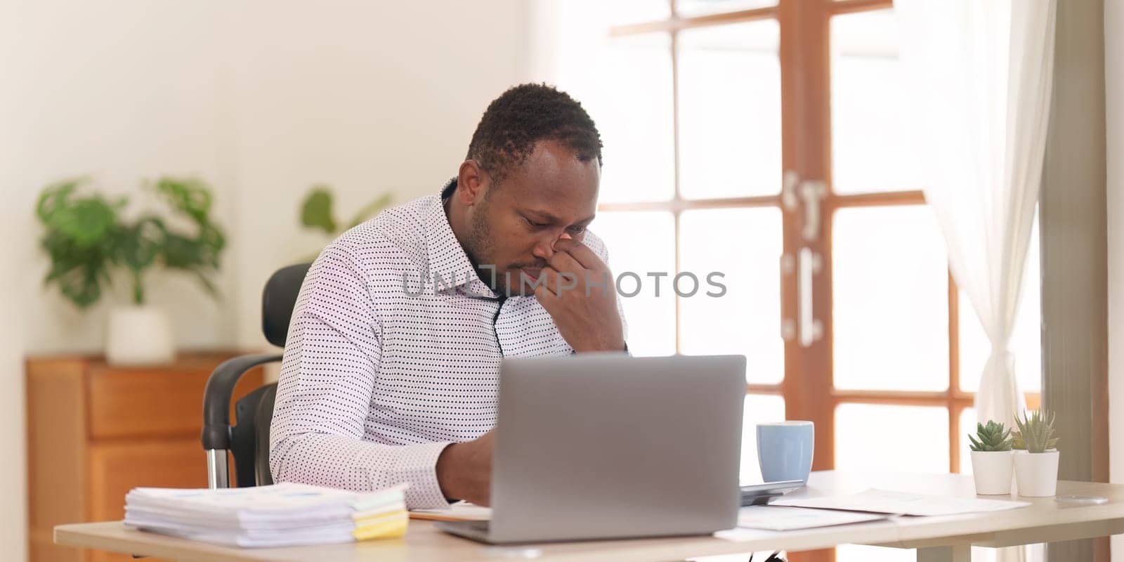 Tired American African business man in stress works with many paperwork document. migraine attack. Freelance, work from home.