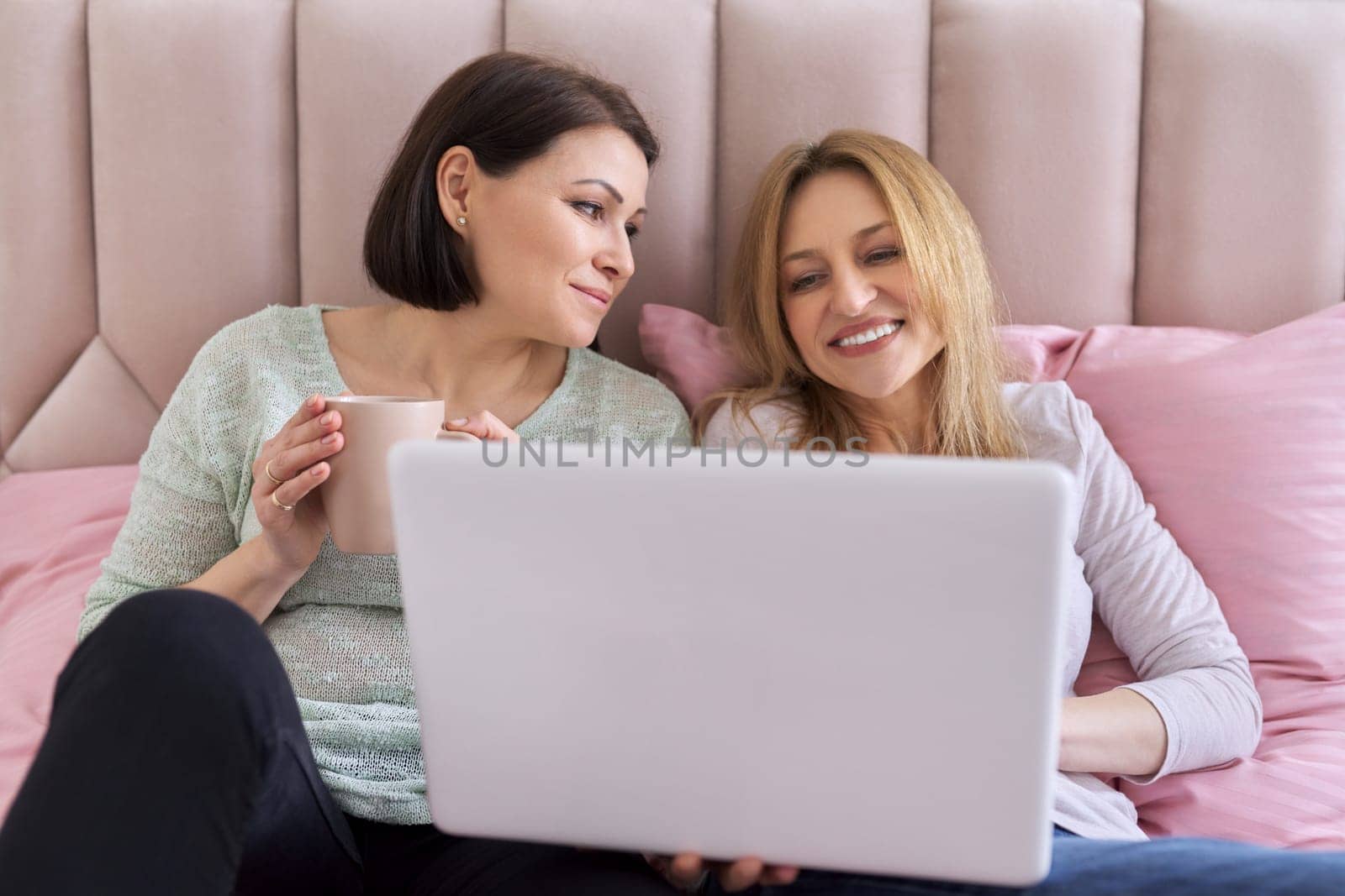 Two middle-aged women sitting at home in bed with laptop, carefully looking at screen, video film, video blog. Rest and leisure, friendship, communication, relationships