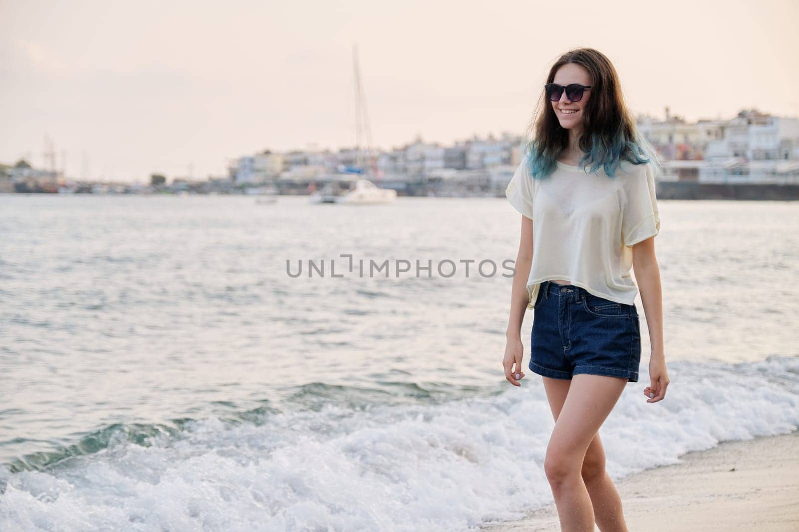 Beautiful teenager girl in sunglasses with dyed blue hair on sea beach, nature sea sky background for copy space. Adolescence, beauty, vacation, weekend concept