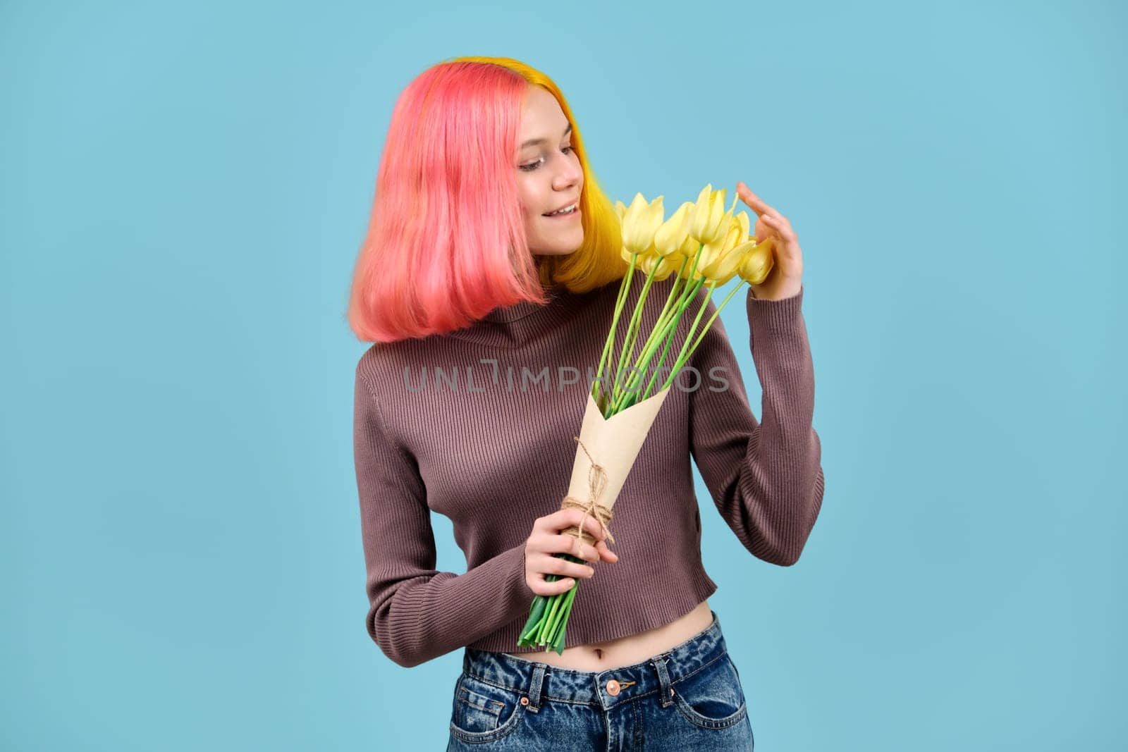 Beautiful smiling trendy teen model with bouquet of yellow tulips on blue studio background. Girl with colored dyed hair, in jeans sweater, enjoying flowers