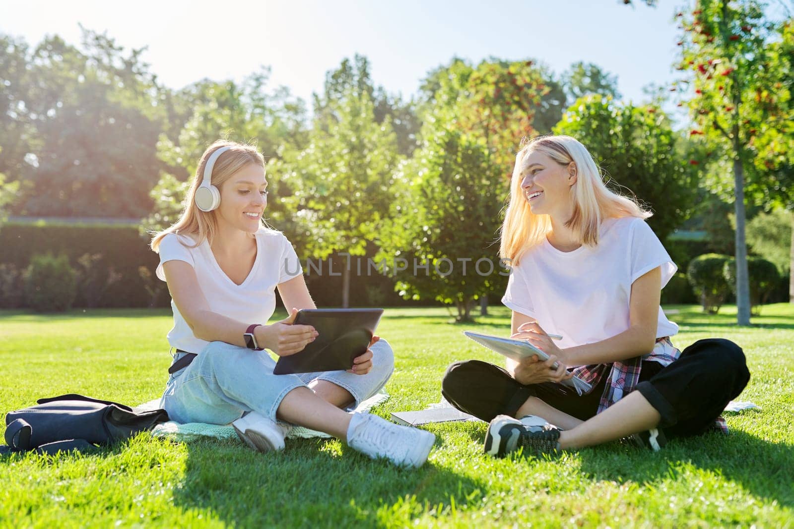 Girls teenage students sitting on green lawn in park with backpack, books, digital tablet, drink bottle of water. University, college, school, education and knowledge, adolescent lifestyle
