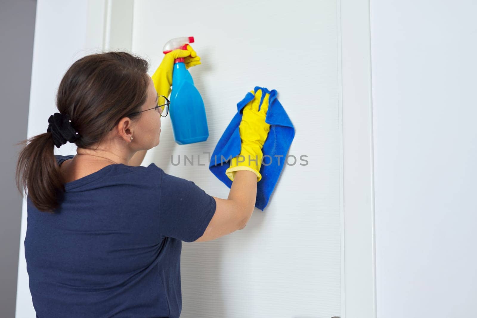 House cleaning, mature woman cleans at home in living room, in yellow gloves with spray, cleans white interior door. Hygiene, purity, cleanliness concept