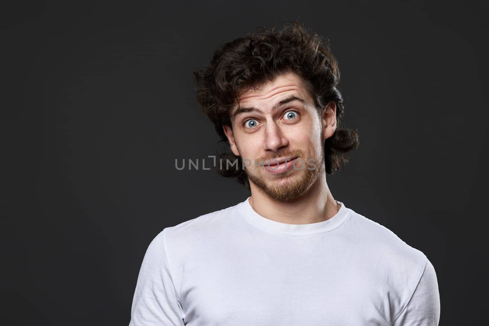 puzzled young bearded man in casual white t-shirt looking at the camera on dark gray background. great idea