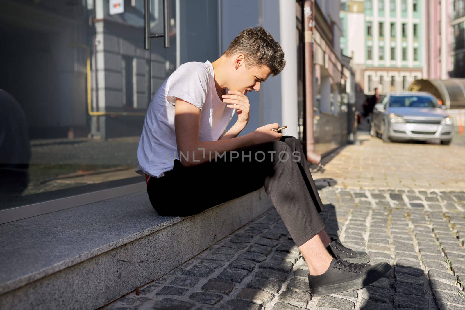 Boy teenager 16,17 years old, using smartphone, sitting on street of summer city, guy reads an online message, browses social networks, types text