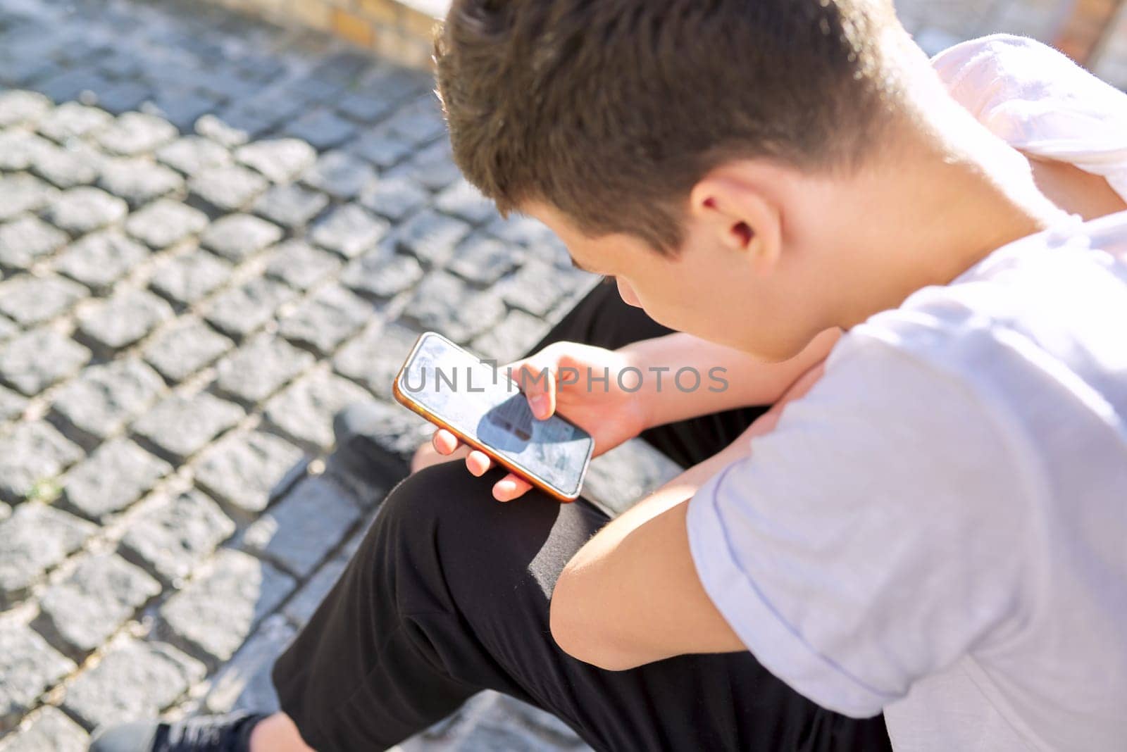 Boy teenager 16,17 years old, using smartphone, sitting on street of summer city. Copy space, guy reads an online message, browses social networks, types text