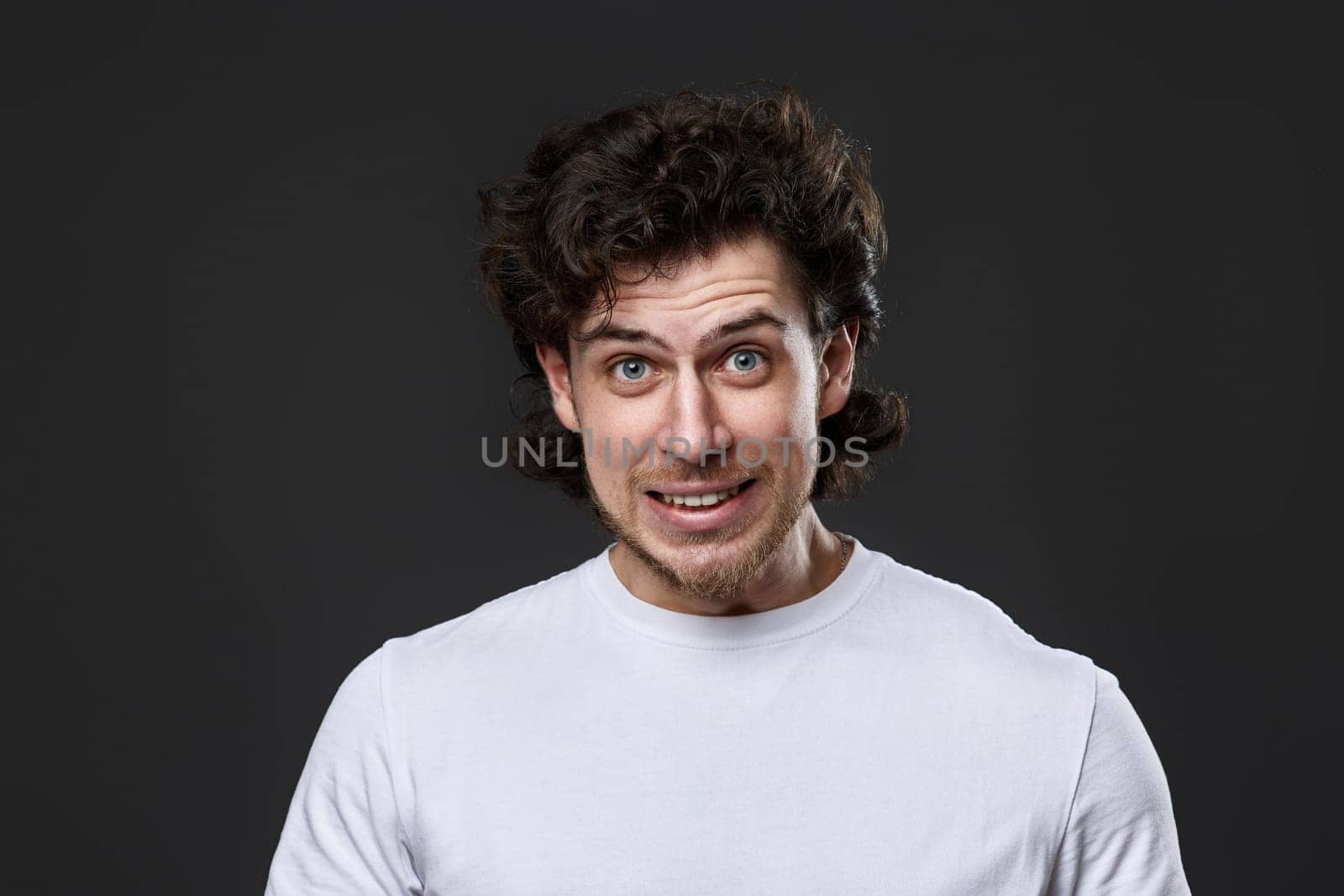 confused young bearded man in casual white t-shirt feels ashamed of terrible mistake on dark gray background. great idea