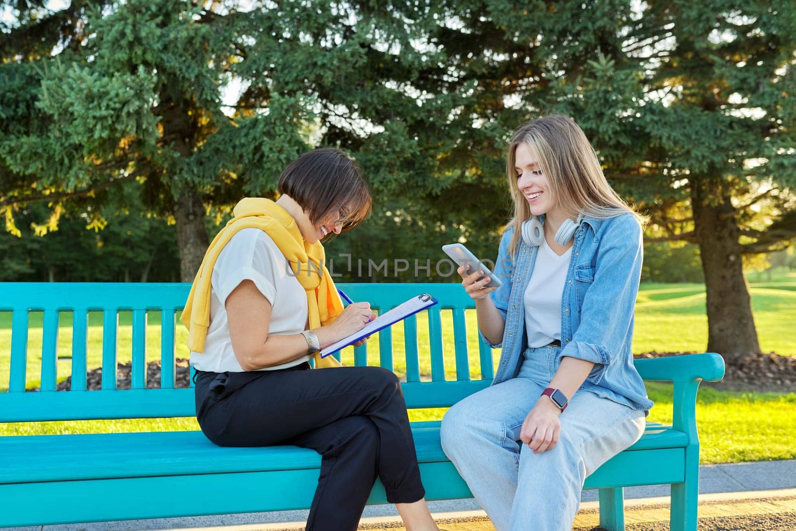 Female social worker, psychologist, journalist, recording interviewing teenage student. Women sitting on bench in the park