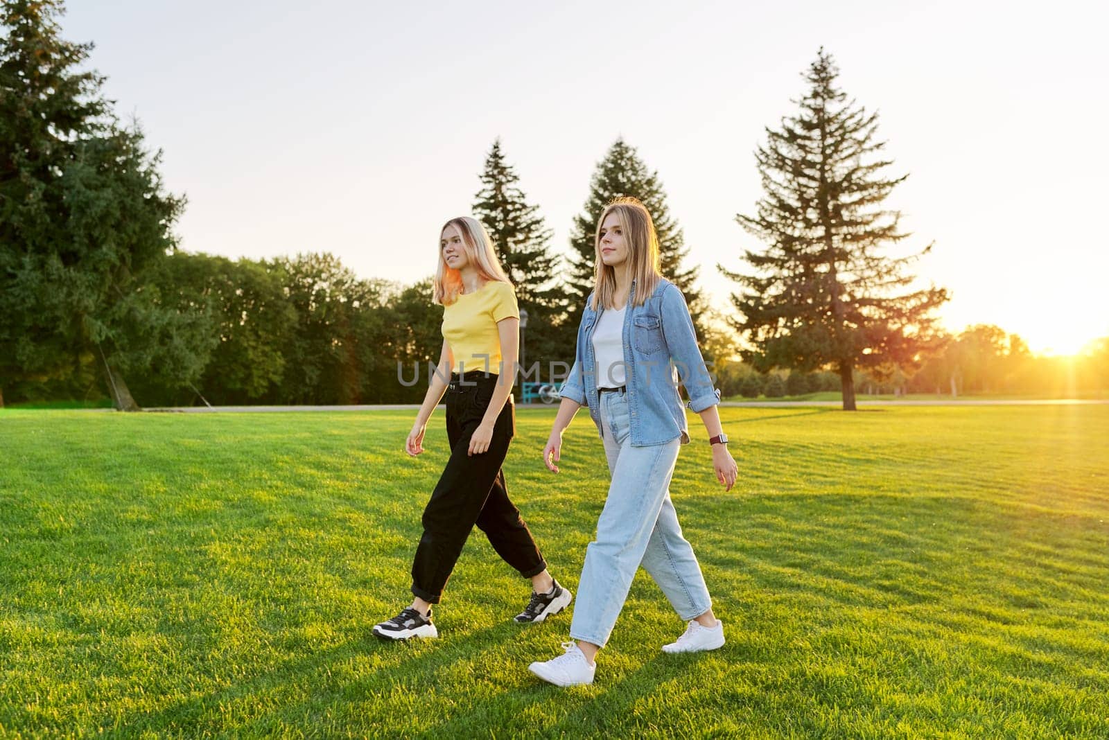 Teen girls walk forward. Two beautiful young women walking together on a green lawn, nature sunset green grass background. Modern youth, adolescence, lifestyle concept