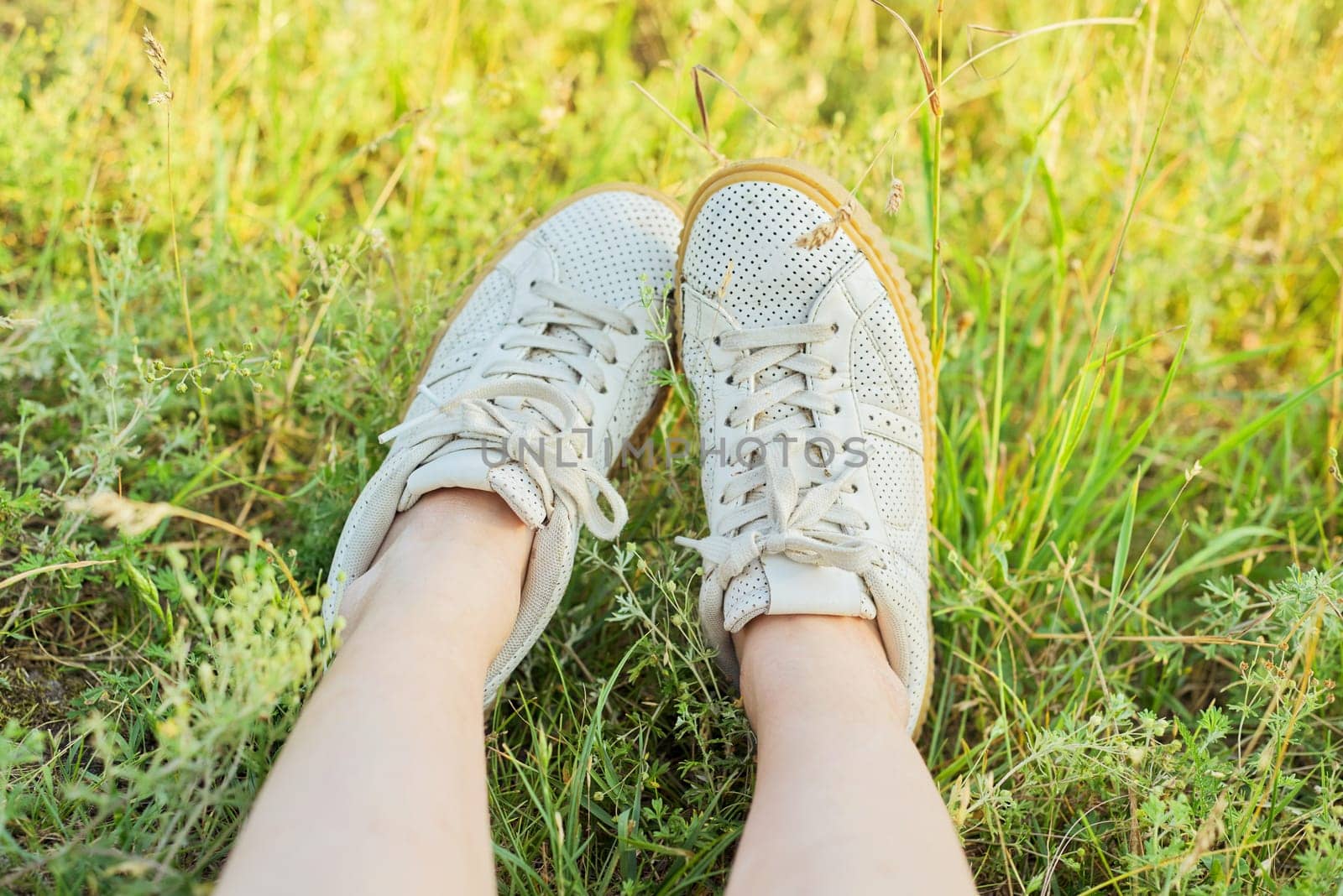 Close-up of female feet in white sneakers in green grass of summer meadow, top view