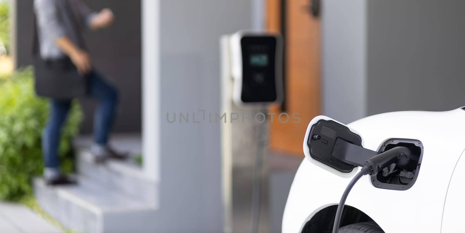 Focus electric car charging at home charging station with blurred progressive man walking in the background. Electric car using renewable clean for eco-friendly concept.