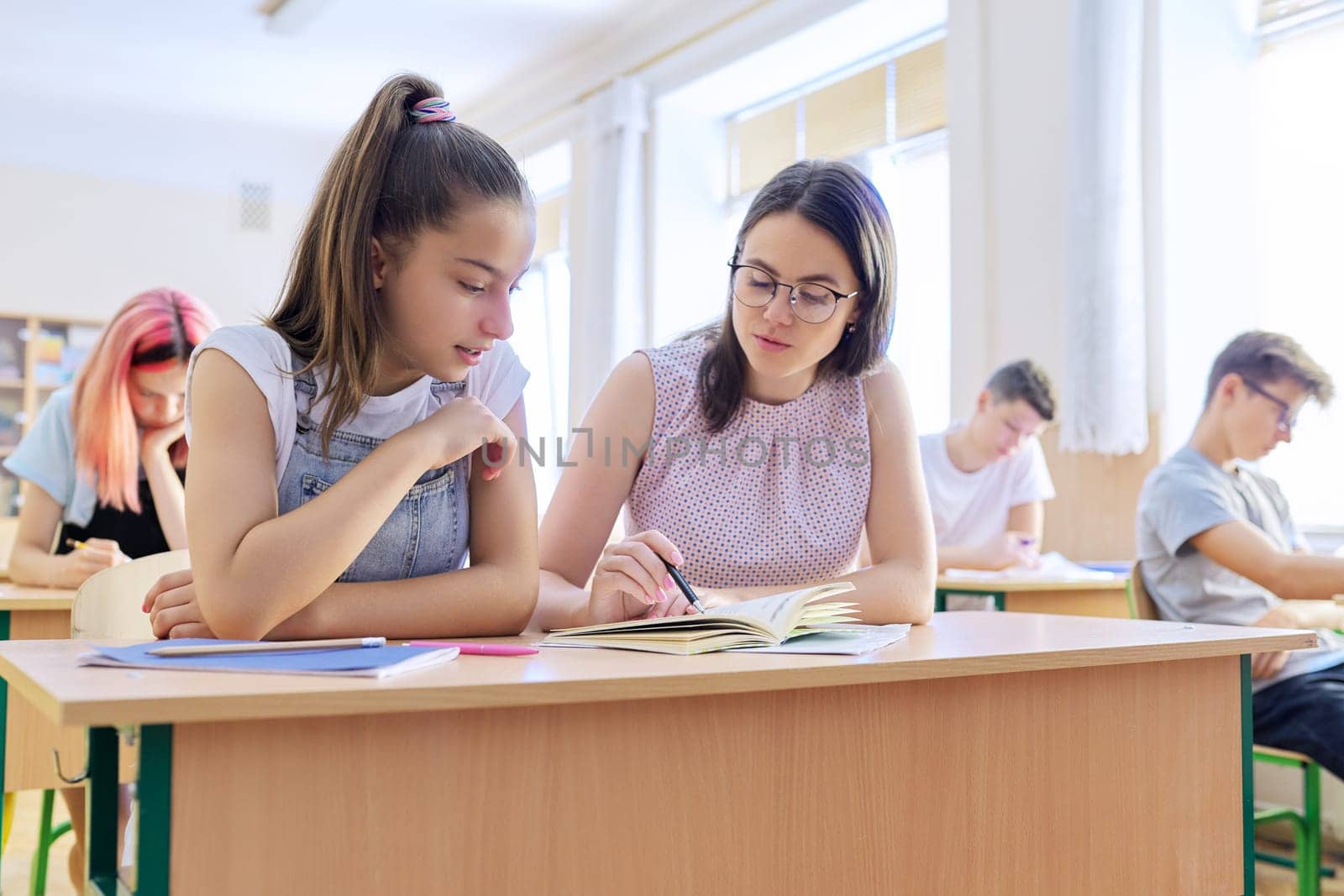 Young woman teacher teaches lesson in classroom of teenage children, teacher sits at desk with student, checks knowledge. Education, school, college, teaching concept