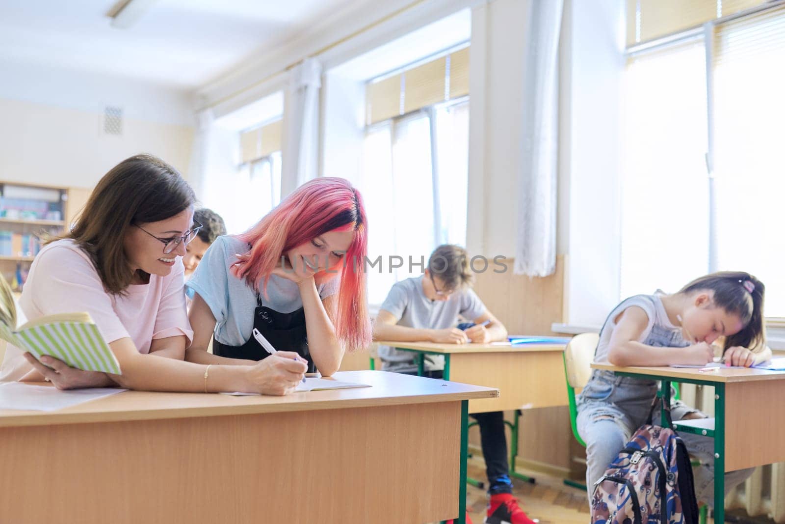 Lesson in class of high school students, female teacher sitting at desk with student by VH-studio