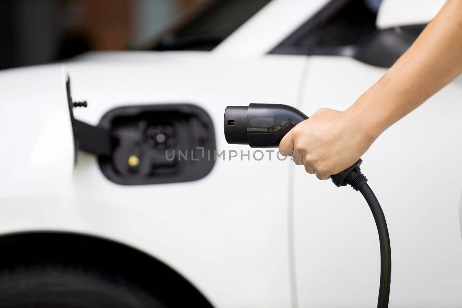 Focus hand holding EV charger plug with blurred background of progressive electric vehicle and socket parking in home garage with electric charging station powered by clean and sustainable energy.