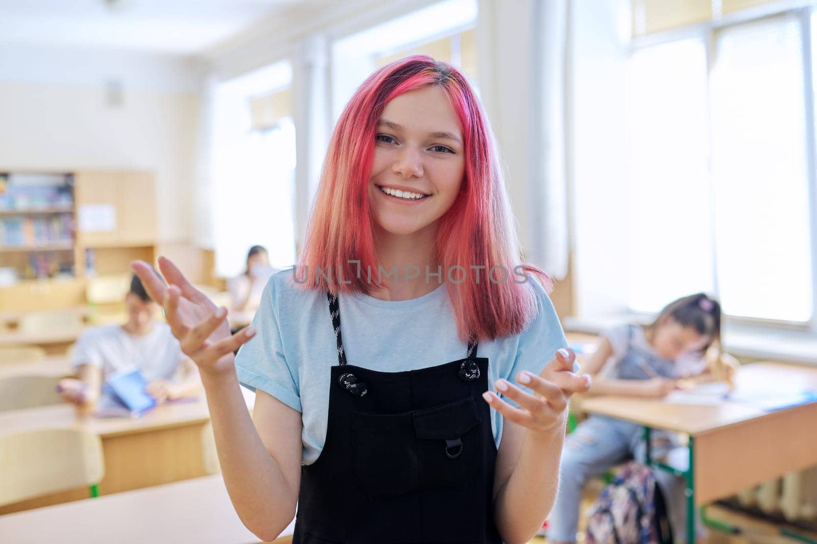 Talking gesturing smiling teenage student at lesson in classroom, trendy beautiful girl looking at camera, background students sitting at desks