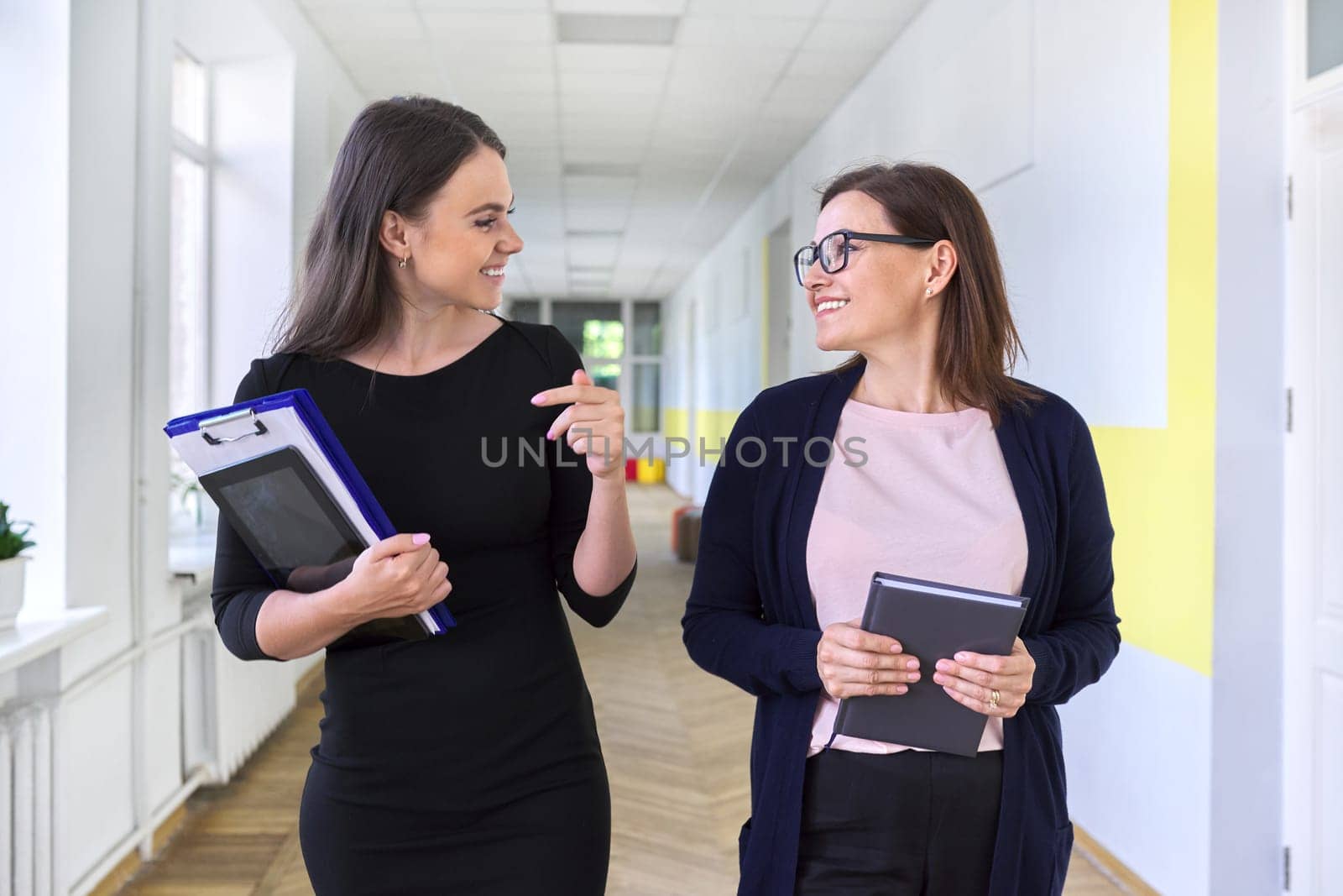 Two colleagues of businesswoman, teacher walking and talking on corridor of office, school. Positive smiling young and middle aged females, business education professions office workers concept