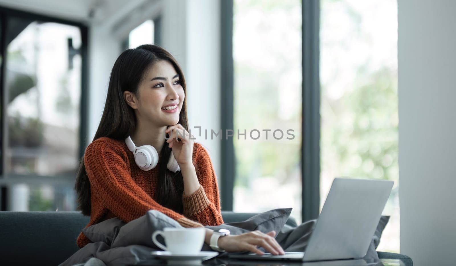 asian young woman listening music with headphone and streaming music from laptop on sofa relaxing at home by wichayada