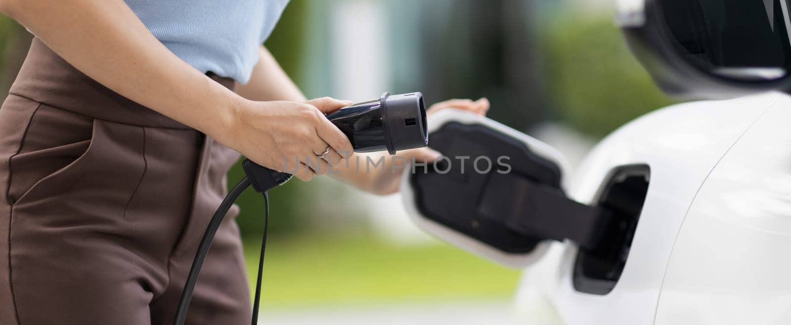 Closeup progressive woman install cable plug to her electric car with home charging station. Concept of the use of electric vehicles in a progressive lifestyle contributes to clean environment.