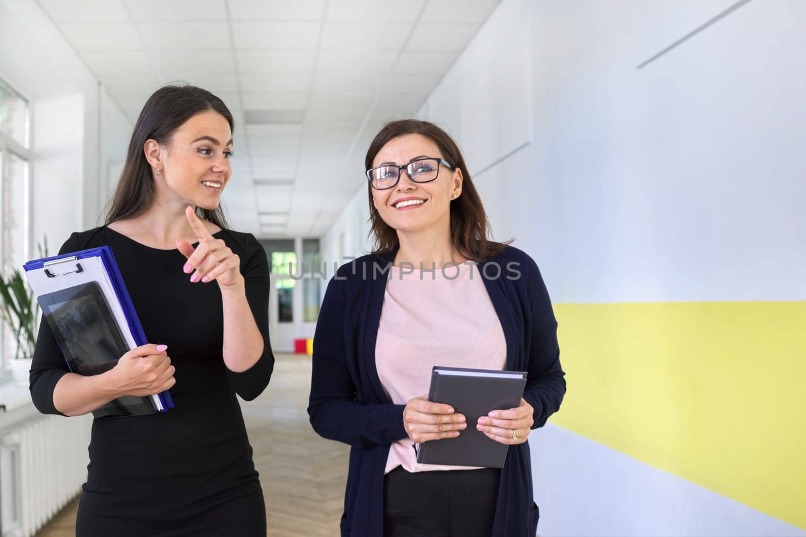 Two colleagues of businesswoman, teacher walking and talking on corridor of office, school. Positive smiling young and middle aged females, business education professions office workers concept