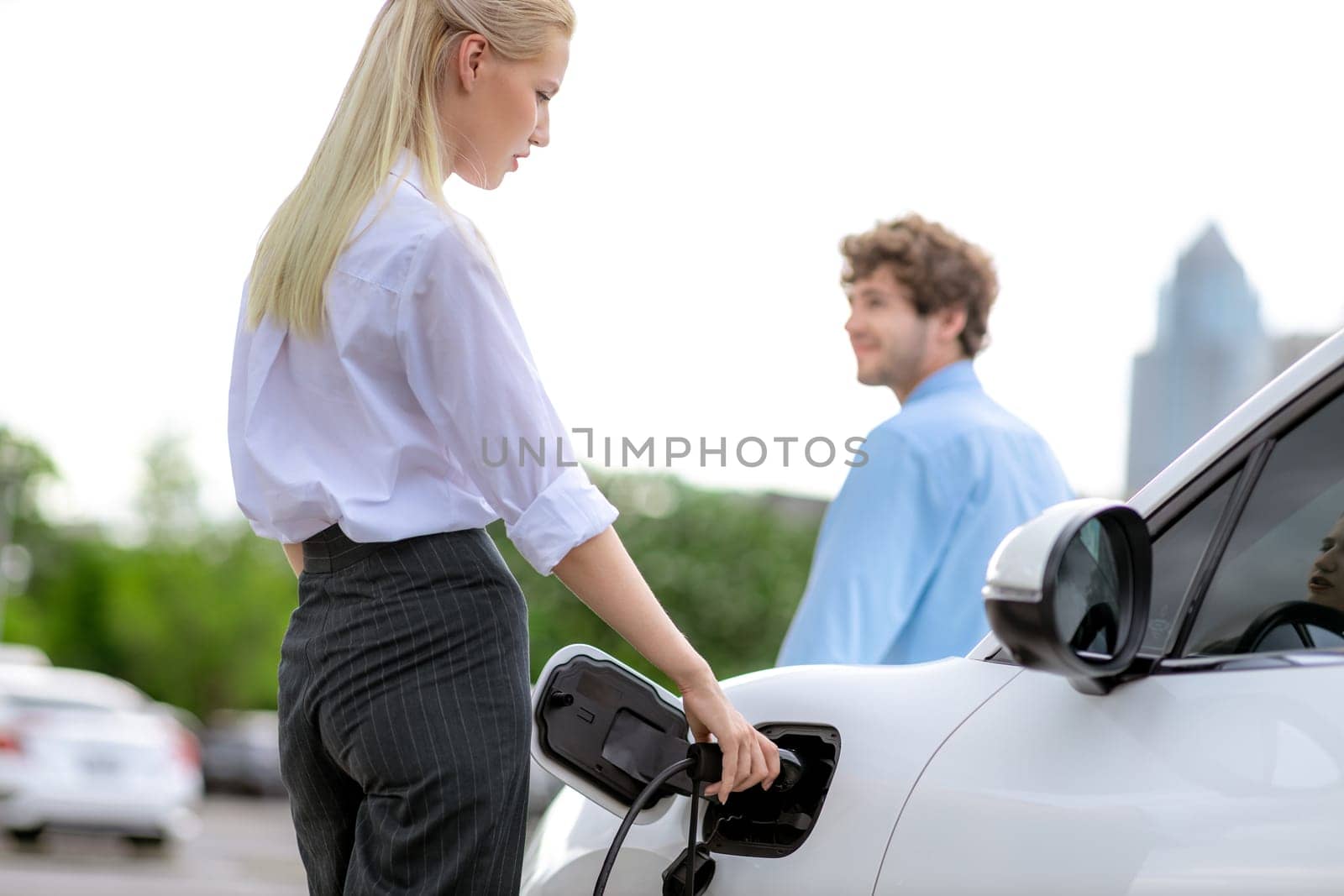 Focus hand insert EV charger to electric vehicle at public charging point in car park with blur business people in backdrop, eco-friendly lifestyle by rechargeable car for progressive concept.