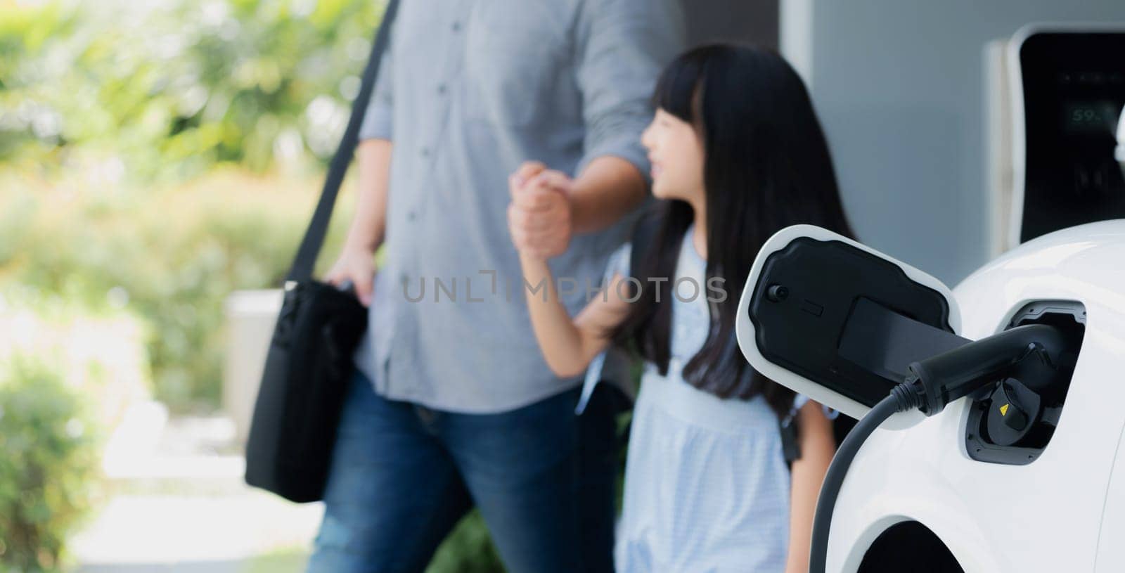 Focus electric car recharging at home charging station with blurred father and daughter walking in background. Progressive green and clean energy vehicle for healthy environment lifestyle concept.