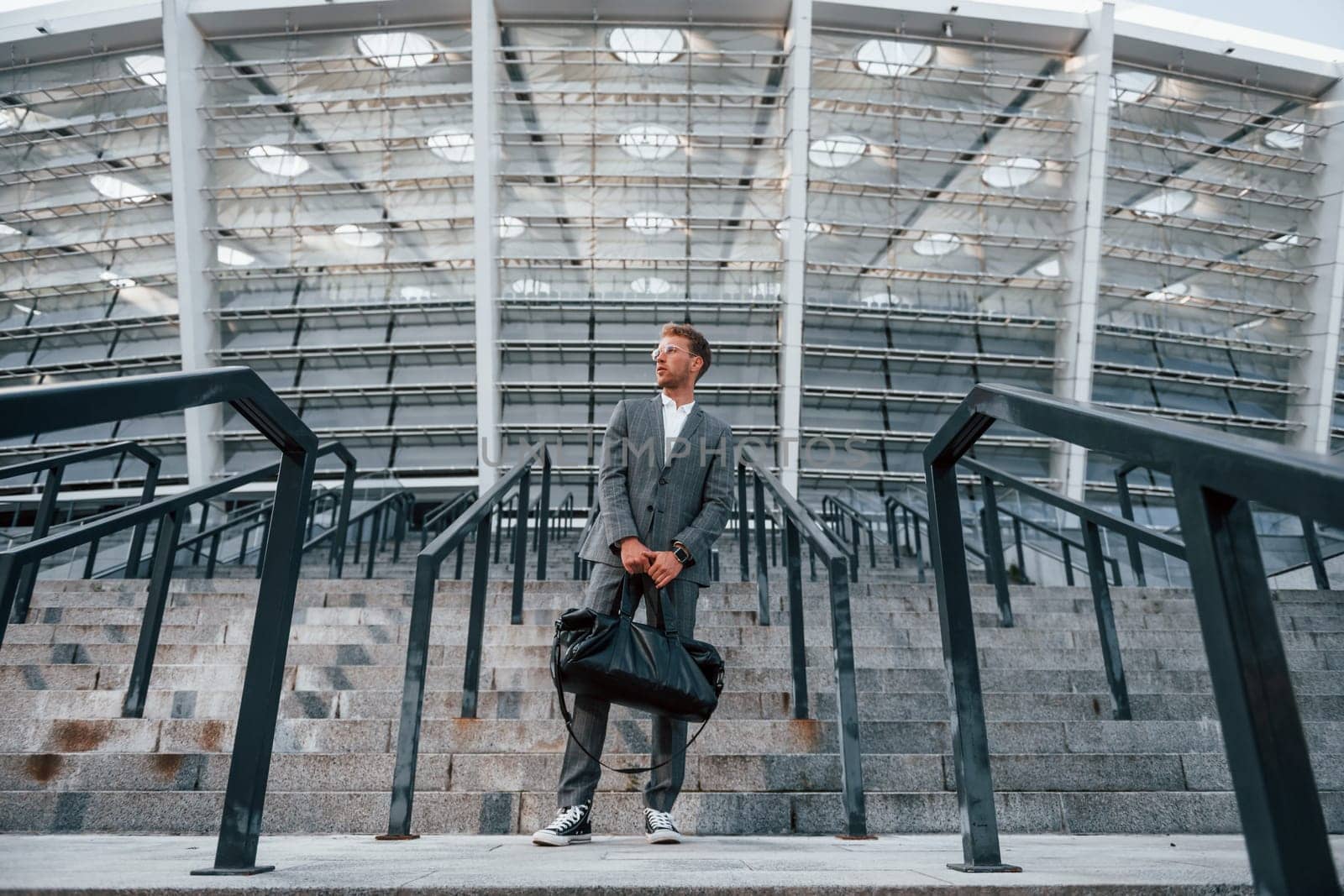 Holds bag in hands. Young businessman in grey formal wear is outdoors in the city.