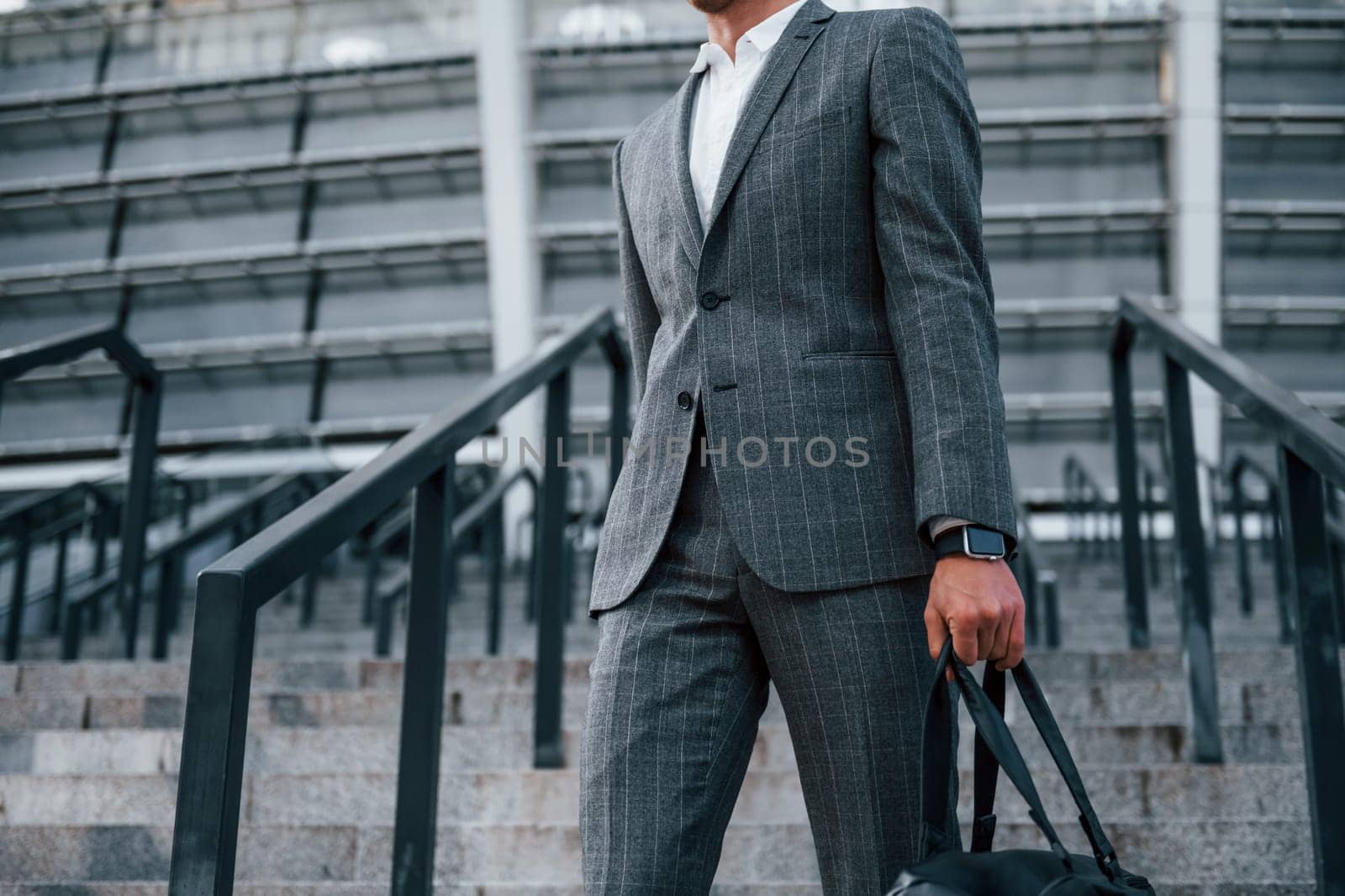 Holds bag in hands. Young businessman in grey formal wear is outdoors in the city.