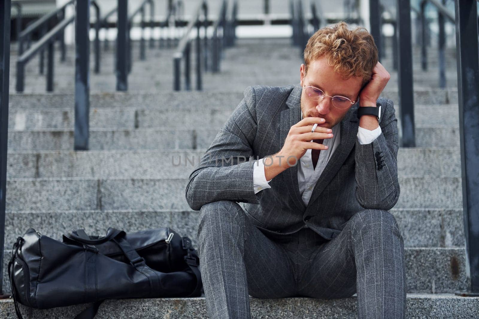 Smoking cigarette. Young businessman in grey formal wear is outdoors in the city by Standret