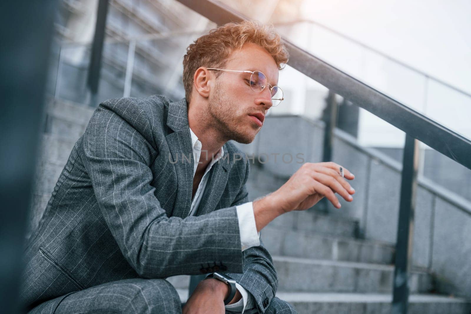 Smoking cigarette. Young businessman in grey formal wear is outdoors in the city.