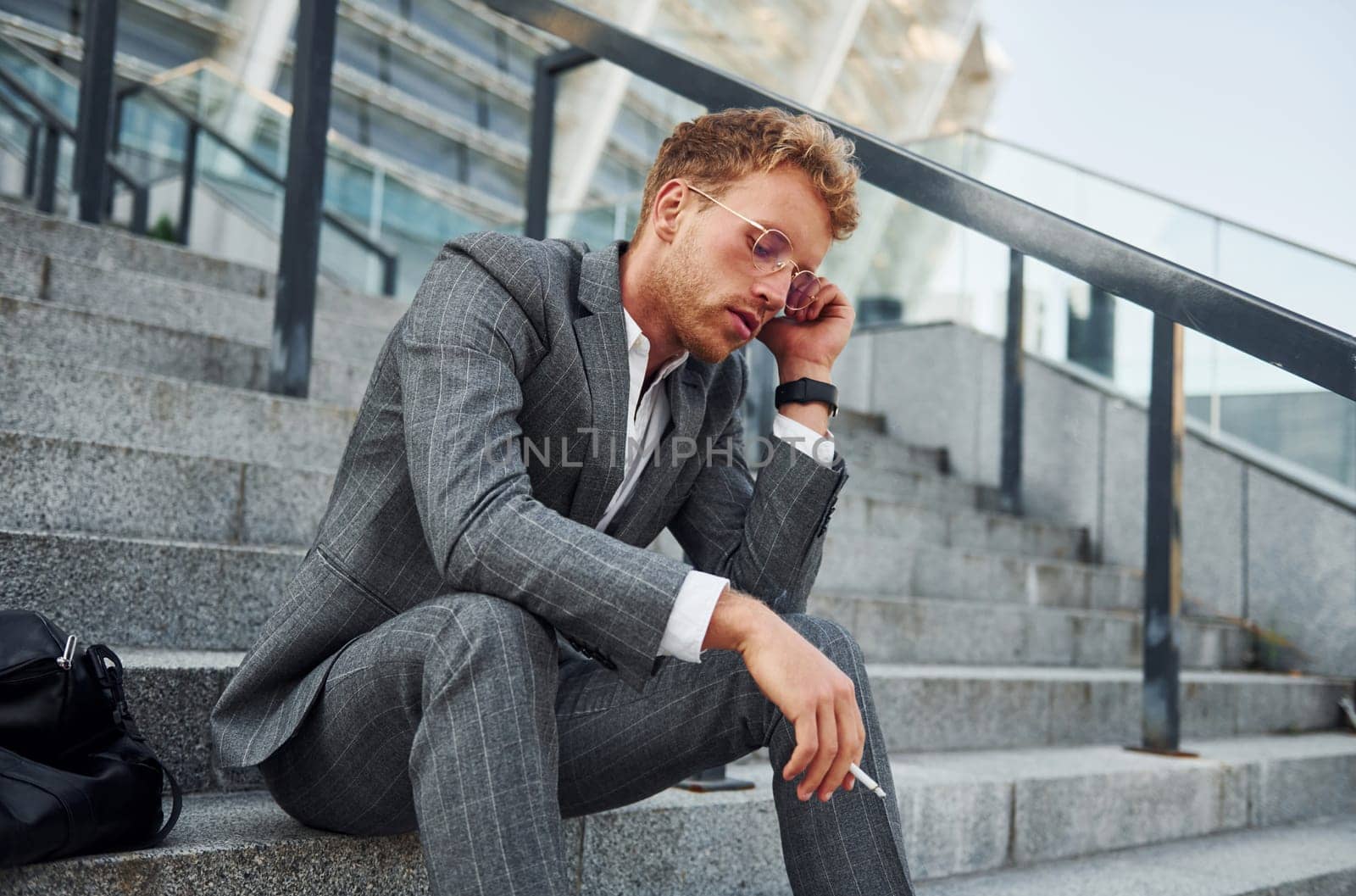 Smoking cigarette. Young businessman in grey formal wear is outdoors in the city.