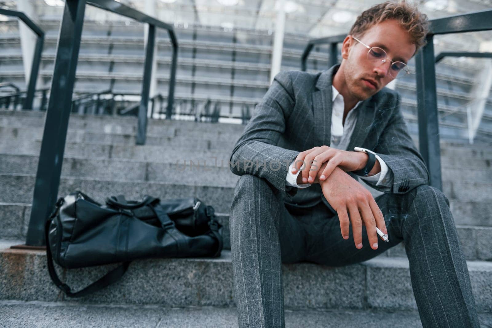 Smoking cigarette. Young businessman in grey formal wear is outdoors in the city.