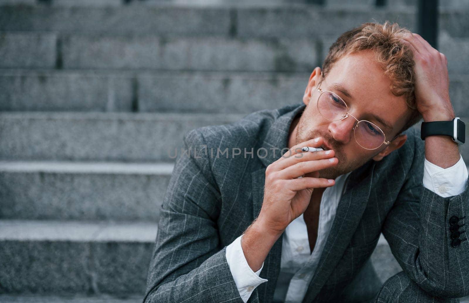 Takes a break and smokes a cigarette. Young businessman in grey formal wear is outdoors in the city.