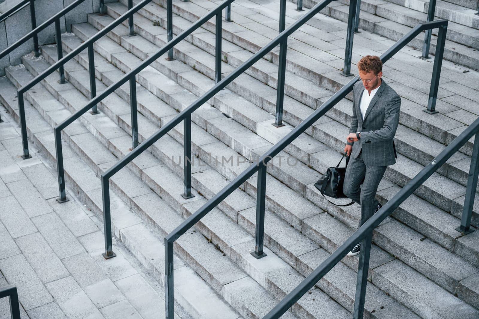 Holds bag in hands. Young businessman in grey formal wear is outdoors in the city by Standret