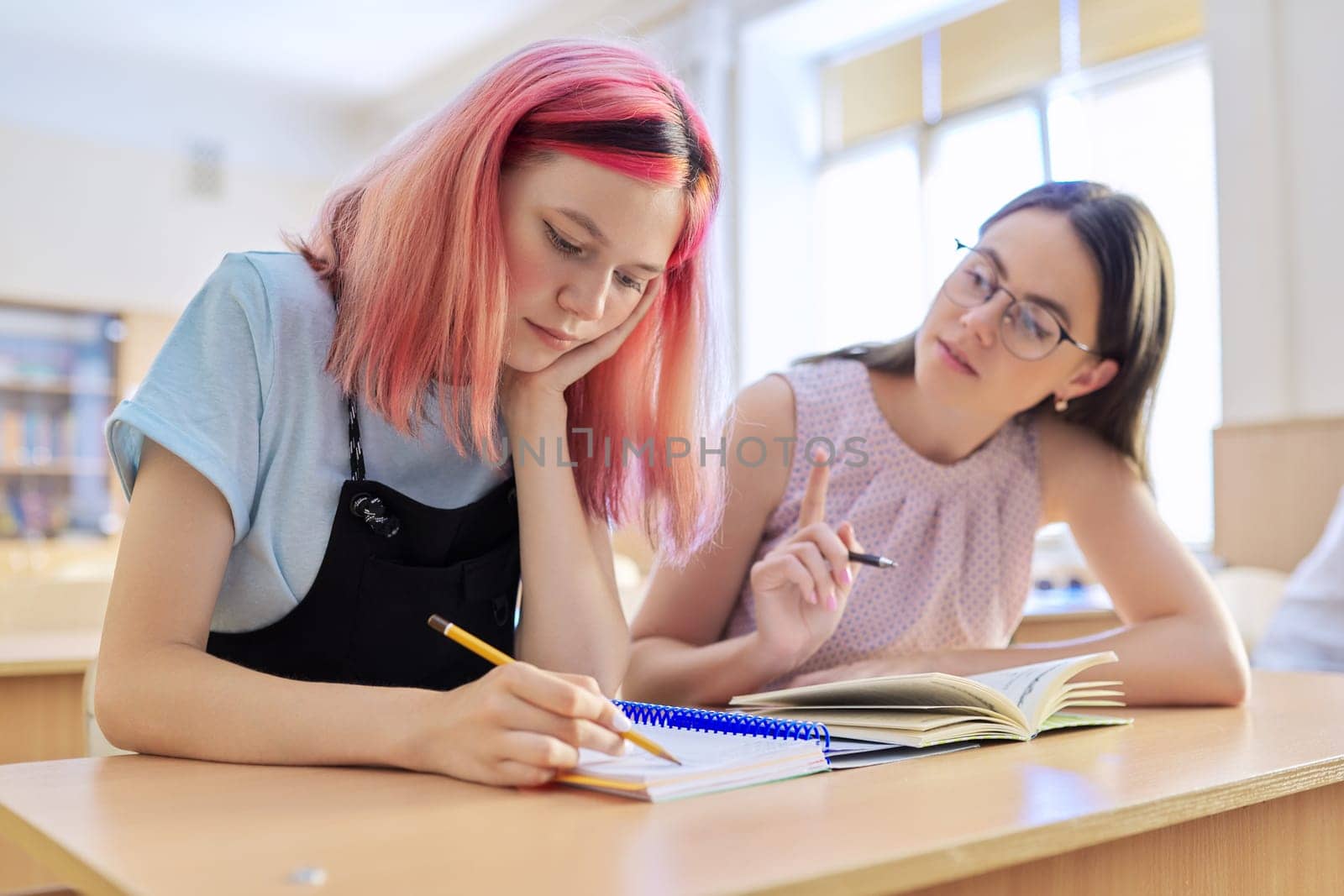 Young woman teacher teaches lesson in classroom of teenage children, teacher sits at desk with student, checks knowledge. Education, school, college, teaching concept