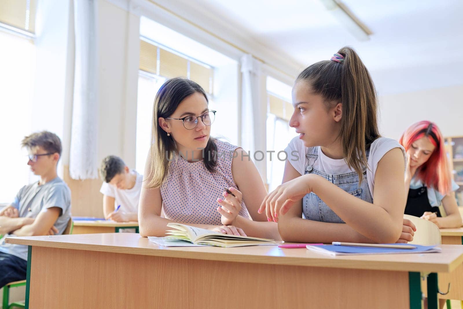 Young woman teacher teaches lesson in classroom of teenage children, teacher sits at desk with student, checks knowledge. Education, school, college, teaching concept