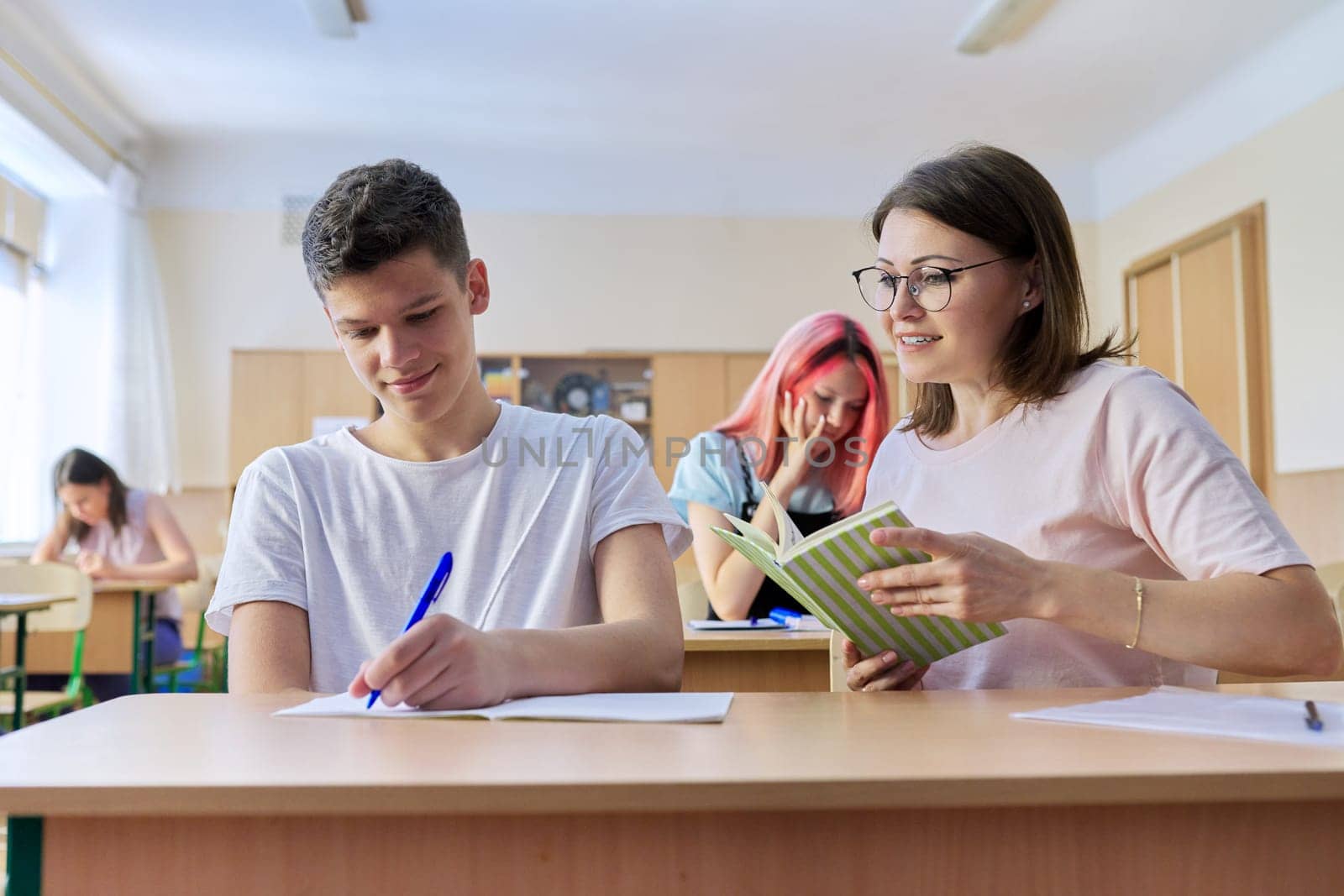 Lesson in the class of high school students, female teacher sitting at desk with male student teenager.