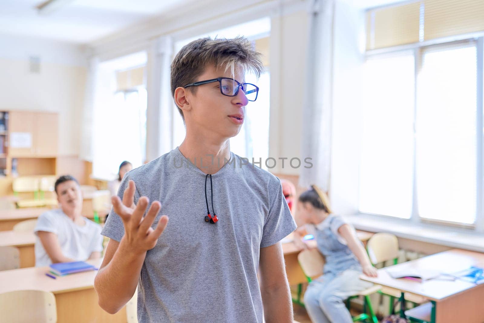 Smart teenager student male taking exam, answers lesson, talking looking at camera, classroom with study students background. High school, education, youth concept